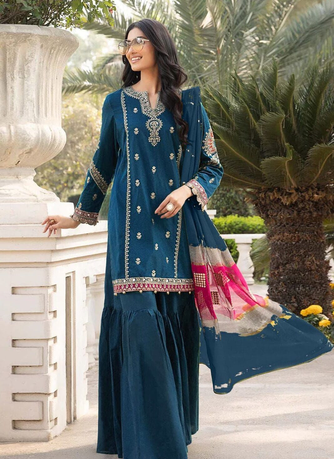 Woman in a teal traditional outfit with pink accents standing outdoors.